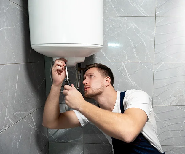 Plombier avec tournevis réparant un chauffe-eau mural blanc sur carrelage gris à Saint-Pathus près de Meaux en Seine-et-Marne 77